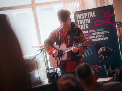 A young man singing and playing guitar. There is an Inspire Youth Arts banner behind him.