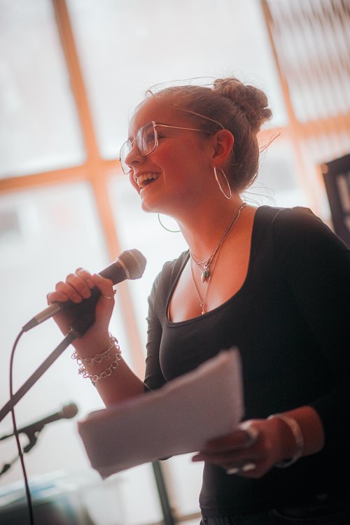 A young woman wearing glasses smiling and singing into a microphone