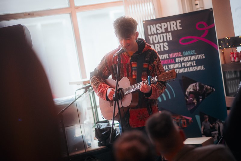 A young man singing and playing guitar. There is an Inspire Youth Arts banner behind him.
