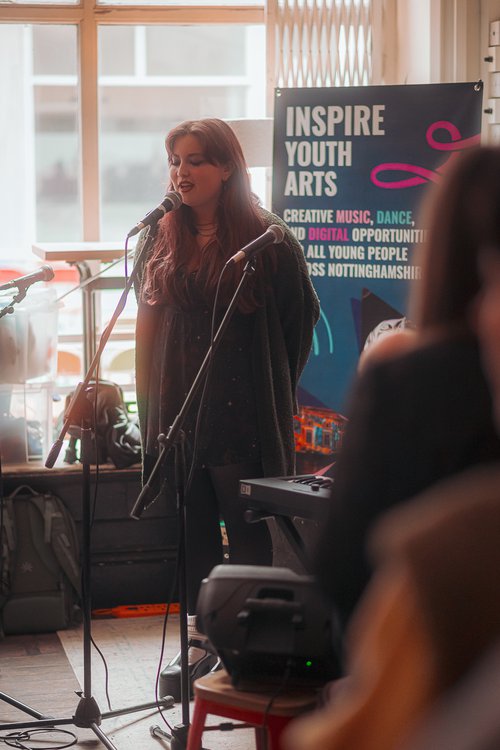 A woman with long hair standing and singing into a microphone. There is a banner that says Inspire Youth Arts and a window behind her.