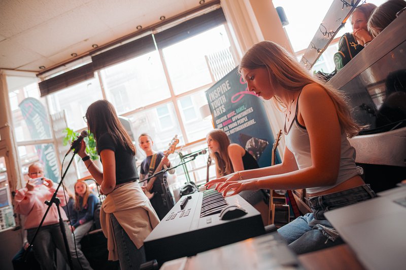 An image from side on of a band. There is a woman on the right playing keys, a woman at the front singing into a microphone and a drummer and bass guitarist in the background.