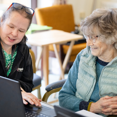 A tutor sits with an older lady during a digital skills course. The tutor points at a laptop screen.