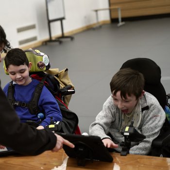 A male music tutor sharing a digital tablet with two young boys, who are smiling and engaged as they look at the screen together.