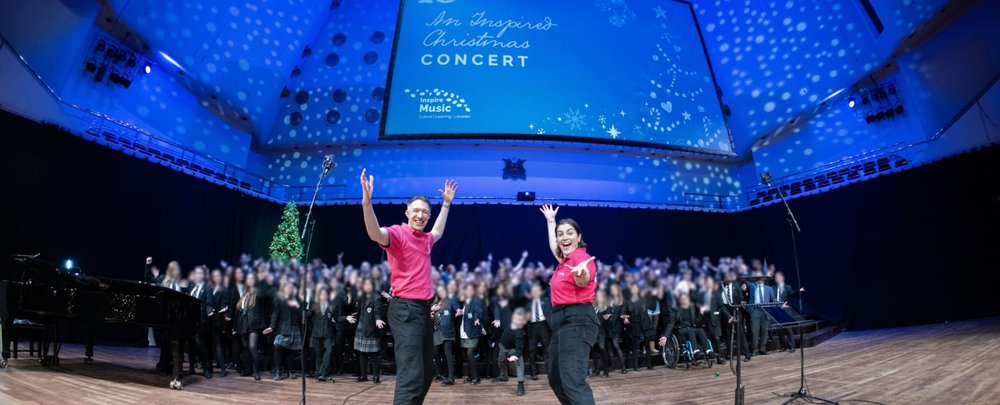 A choir on stage at christmas with two National Youth Choir workshop hosts on stage
