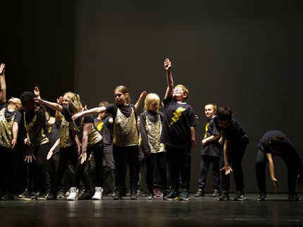 12 young dancers on a black stage, with their arms out at different angles. They are wearing black and gold costumes