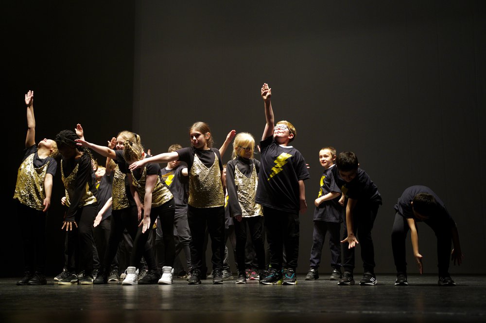 12 young dancers on a black stage, with their arms out at different angles. They are wearing black and gold costumes