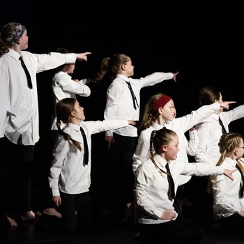 8 young people wearing white shirts and black ties on stage pointing to the right