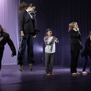 Seven young dancers wearing suits jumping in the air on stage