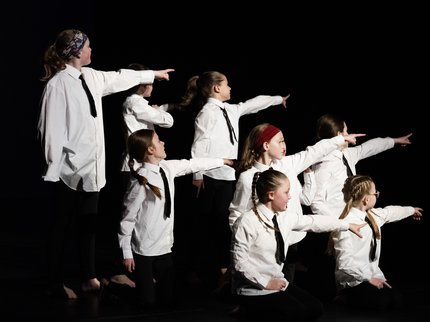 8 young people wearing white shirts and black ties on stage pointing to the right