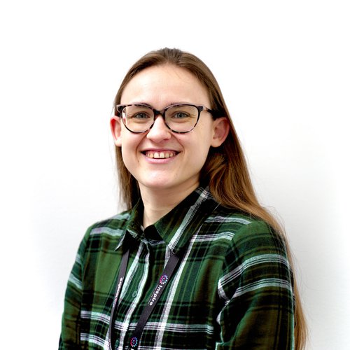 Female with long brown hair, glack framed glasses, smiling to camera. Wearing a green and white check shirt standing against a white background.