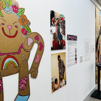 The exhibition wall featuring a large cardboard puppet and photographs of the artist and author in residence
