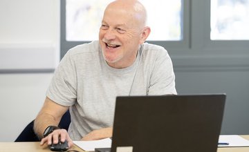 A middle aged man wearing a light grey t-shirt sits at a computer, holding a mouse whilst smiling. The man is white with a bald head, moustache and goatee beard.
