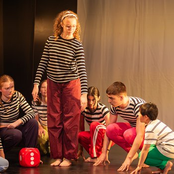 One dancer standing, 8 others crouched down. All wearing black and white stripe tops and bright colour bottoms. There is a large red and white dice on the floor
