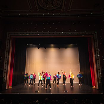 Dances on a large stage, with red curtains, a disco ball. Dancers in bright colours, jumping