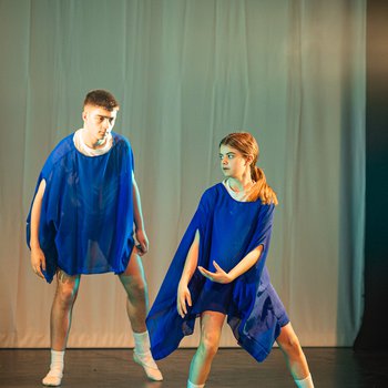 Two dancers in floaty blue tops, black shorts and ballet shoes lunging to their left