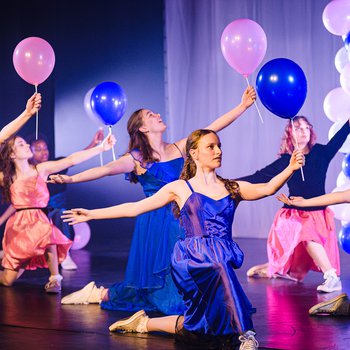 6 dancers, all wearing different colour prom dresses, holding pink blue and purple balloons on sticks. Dancers are kneeling