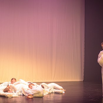 6 dancers with pillows laying in a clump. One dancer to their right holiday a pillow and jumping with one arm and leg up