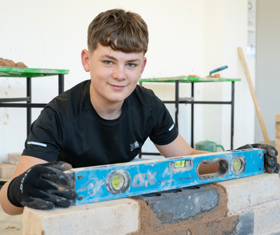 A construction student smiles at the camera whilst checking that a brick wall is level