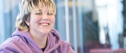 A student in a light purple hoodie is using their laptop and smiling at the camera
