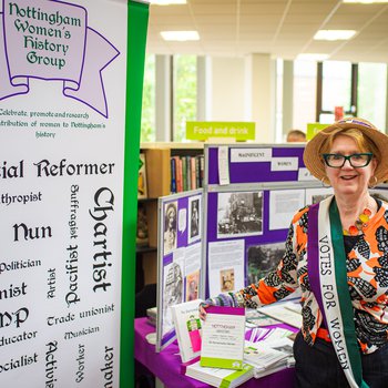 A female stallholder from Nottingham Women's History Group, stood with a banner and display and wearing a 'Votes for Women' sash.