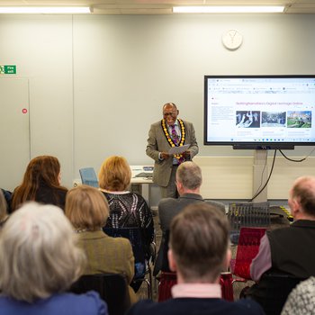 The Chair of Nottinghamshire County Council introducing Inspire's Nottinghamshire digital heritage platform (shown on a large TV screen) to a seated audience.