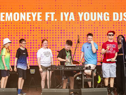 A group of young people on a stage with an orange background, the boy in the middle is DJing