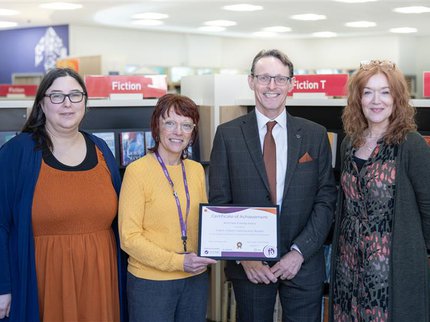 Group of 4 smiling people, 3 female and 1 male in a library setting receiving a certificate