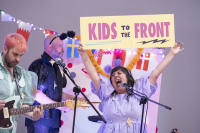 Three performers on stage holding sign 'Kids to the Front#