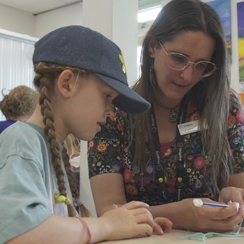 A child in a denim baseball cap watches a lady with long brown hair and glasses demonstrate how to knit
