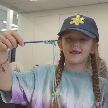 A child in a denim baseball cap holds up a knitting needle with several rows of knitting