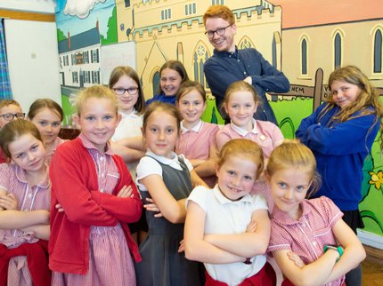 A group of children in school uniform stand with two vocal coaches