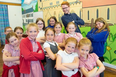 A group of children in school uniform stand with two vocal coaches