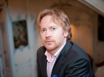 A photograph of poet Paul Farley - a man with light coloured hair and a goatee beard, wearing a grey suit.