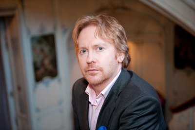A photograph of poet Paul Farley - a man with light coloured hair and a goatee beard, wearing a grey suit.