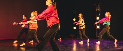 Female dancers in brightly coloured jackets on stage with one arm raised.