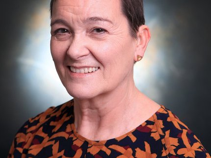 Woman with short brown hair, smiling and wearing small gold triangular earrings and a patterned brown and orange top.