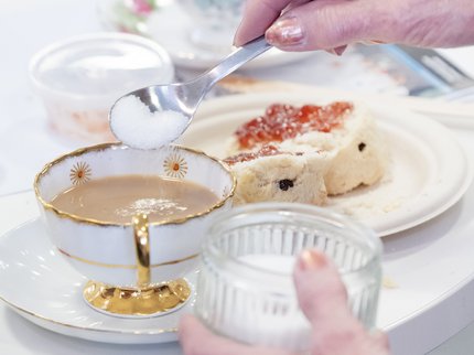 Photograph of a cup of tea with someone adding a spoon of sugar with a scone on a plate next to the cup and saucer.