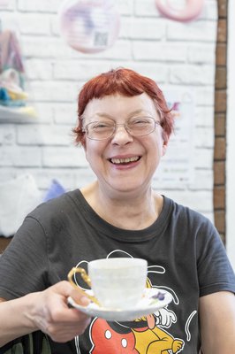A person with short red hair is holding a cup of tea and saucer and smiling at the camera.