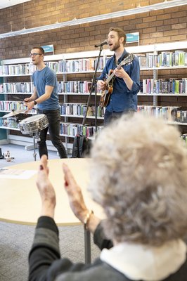Man playing a guitar and a man playing the drums with a woman clapping in the foreground