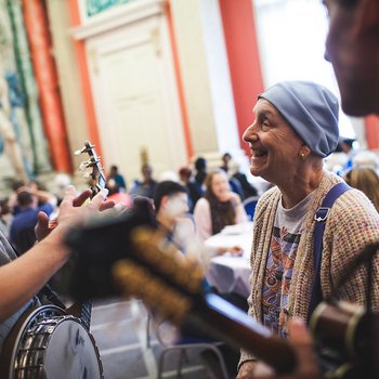 A busy room, in the foreground a lady is taking to a musician