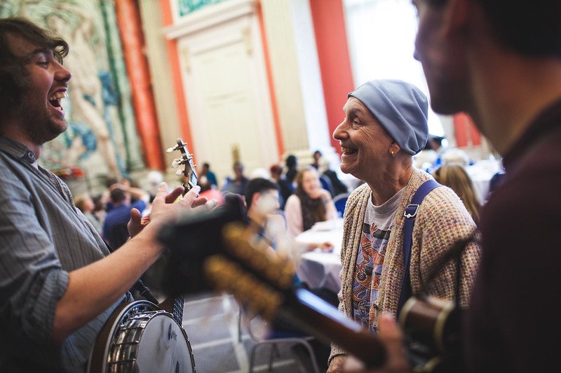 An elderly woman is smiling broadly at a smiley musician who is playing the guitar.
