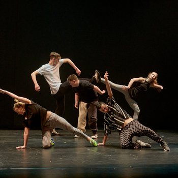 Five young dancers dressed in black and white performing on stage with their legs outstretched.