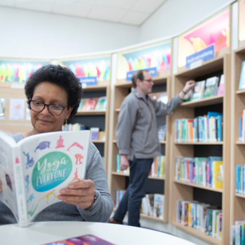 Person sat reading a book while another reader browses the shelves behind them