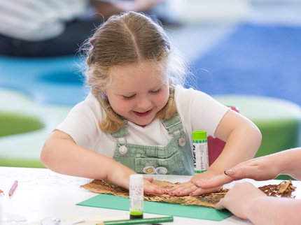 A young white girl with blonde plaits is doing arts and crafts in a library