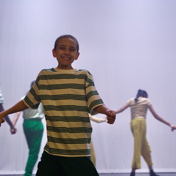 Young dancers holding hands in a circle facing out, wearing green, yellow and pink clothes. One boy at the front smiling to camera
