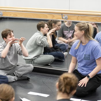 7 dancers sat in a circle smiling. One has their hands to their face describing something. They are sat on a  black floor with a mirror behind them with the groups reflection in.