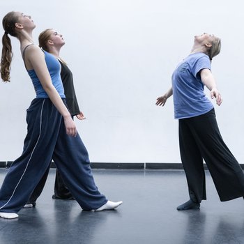 3 dancers al in black trsouers, one in a purple top, one in a blue top and one in a black top, looking up to the sky. Black dance floor and white walls.