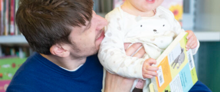 a man in a blue jumper is smiling and looking at the toddler he's holding. toddler has ginger hair and is holding a picture book