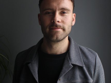 man smiling in front of a dim lit background, he has a stubbled beard and is wearing a grey jacket
