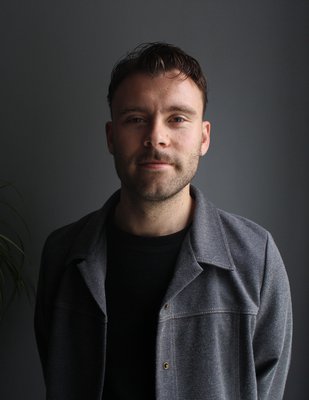 man smiling in front of a dim lit background, he has a stubbled beard and is wearing a grey jacket